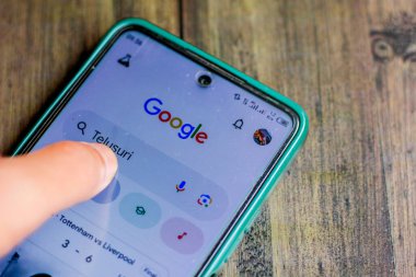Endonezya, Solo - 26 Aralık 2024: İnsan parmağı akıllı telefon ekranına dokunuyor Google uygulama menüsü ahşap masa arkasında izole edildi