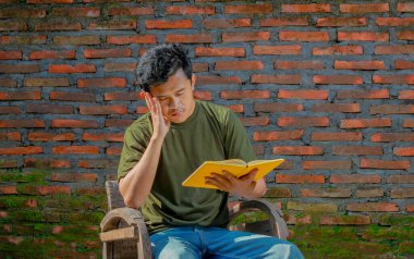 Asyalı bir adam sandalyede oturmuş tuğla duvarlı bir kitap okuyor.