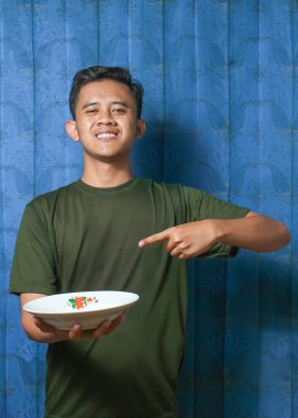 Asyalı bir adam cam levhayı tutarken, mavi perde arkasını parmağıyla işaret ediyor.