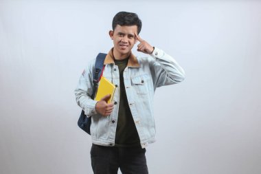 Kot ceket giyen Asyalı bir adam çanta ve kitap taşıyor. Ciddi bir düşünce işareti veriyor. Parmağını kafasına doğrultuyor ve çok düşünüyor.