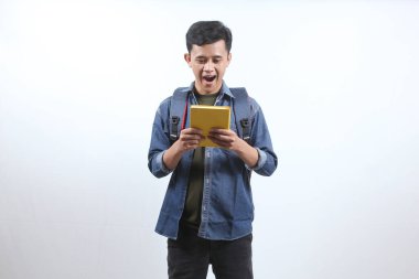 Koyu mavi kot gömlek giyen Asyalı bir adam sırt çantası taşıyor ve beyaz arka planda izole edilmiş bir şekilde hediye kitabına mutlu bir yüz ifadesi alıyor. eğitim kavramı
