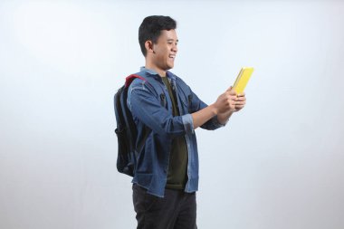 Koyu mavi kot gömlek giyen Asyalı bir adam sırt çantası taşıyor ve beyaz arka planda izole edilmiş bir şekilde hediye kitabına mutlu bir yüz ifadesi alıyor. eğitim kavramı