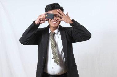 Asyalı işadamı, bir gözünü kapatıp, beyaz arka planda izole edilmiş üst düzey kredi kartı hizmetini destekleyen bir kredi kartını elinde tutarak gizlice gözetlemesine şaşırdı.