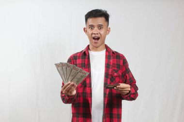 Şok olmuş Asyalı adam kırmızı ekose gömlek giyiyor beyaz tişört, elinde fan dolarları ve kredi kartı var.