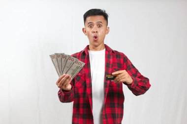 Şok olmuş Asyalı adam kırmızı ekose gömlek giyiyor beyaz tişört, elinde fan dolarları ve kredi kartı var.
