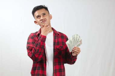 Asyalı genç adam, kırmızı ekose gömlek giyiyor. Elinde bir sürü dolar var. Ciddi düşünürken, beyaz arka planda soyutlanmış.