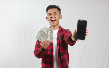 Asyalı adam evde internet üzerinden çalışarak para kazanıyor, elinde siyah ekran akıllı telefon ve birkaç dolar fatura tutuyor. İş ve finans kavramı.