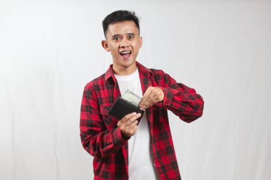 Kareli gömlek giyen Asyalı adam beyaz arka planda izole edilmiş cüzdandan dolar alıyor.