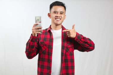 Kareli gömlek giyen Asyalı bir adam beyaz arka planda izole edilmiş 100 dolarlık banknotu işaret ediyor.