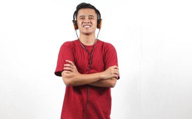 Beyaz arka planda kollarını kavuştururken kulaklıkla müzik dinleyen gülümseyen Asyalı adam.