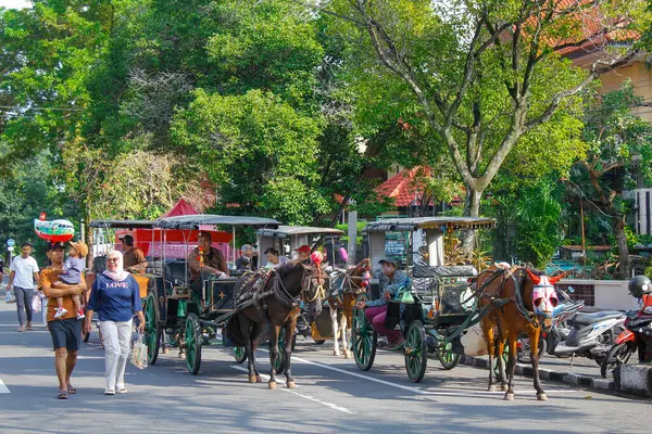 Endonezya, Solo - 1 Haziran 2025: Geleneksel at arabası çizgili elbiseli yaşlı bir adam tarafından kullanılıyor ve yol kenarında durup yolcuları bekliyor.