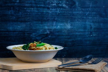 Alüminyum kaşık ve çatalın yanında kesme tahtasının üzerinde yenmeye hazır lezzetli bir tavuklu erişte kasesi.