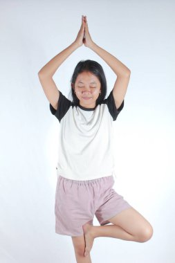 Beyaz tişört ve şort giyen güzel Asyalı kadın portresi ve yoga yaparken mutlu olmak, Vrksasana duruşu, Tree Posure. Beyaz arka planda izole edilmiş.
