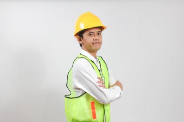 Kendine güvenen genç bir inşaat işçisinin portresi muhteşem bir bina inşa etmek için kollarını kavuşturuyor. Yeşil bir yelek, beyaz gömlek ve beyaz bir arka planda izole edilmiş maden şapkası vardı.