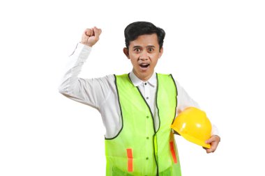Sarı şapkalı, güvenlik yeleği giymiş genç Asyalı inşaat işçisi bir kutlama jesti olarak yumruğunu kaldırıyor. Beyaz bir arkaplanda izole edilmiş.