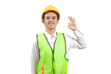 Sarı sapkalı ve neon yeşil güvenlik yeleği giyen gülümseyen Asyalı inşaat işçisi onay işareti veriyor. Beyaz bir arkaplanda izole edilmiş