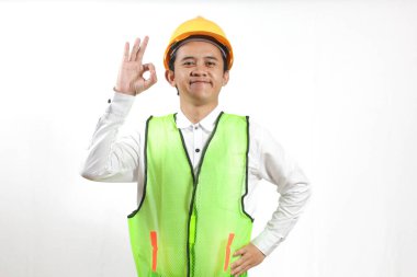 Sarı sapkalı ve neon yeşil güvenlik yeleği giyen gülümseyen Asyalı inşaat işçisi onay işareti veriyor. Beyaz bir arkaplanda izole edilmiş