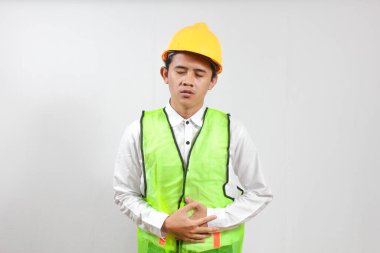 Sarı kasket ve güvenlik yeleği giyen genç inşaat işçisi mide ağrısı çekiyor ve endişeli bir yüz ifadesiyle karnını tutuyor. Beyaz arkaplanda izole.