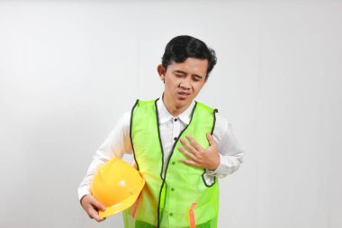 İnşaat yeleği giyen endişeli bir adam göğsünde ağrı hissediyor ve eliyle göğsünü tutuyor. Bu da iş yerinde olası bir kalp krizi ya da başka bir sağlık sorunu olduğunu gösteriyor..