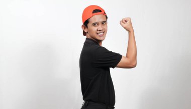 Kaslarını esneten, siyah polo tişörtlü ve turuncu şapkalı beyaz arka planda gülümseyen kendine güvenen genç adam.