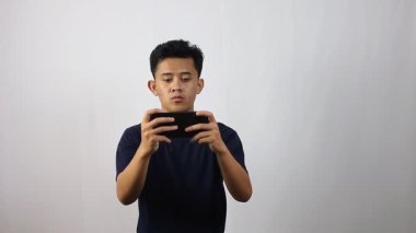 Genç Asyalı adam, beyaz arka planda izole edilmiş akıllı telefonuyla oyun oynarken çok şaşırdı.