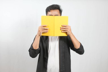 Gözlüklü genç adam sarı bir kitabın arkasına saklanıyor. Beyaz arka planlı stüdyo çekimi. Okuma, öğrenme, eğitim ve mahremiyet kavramı.