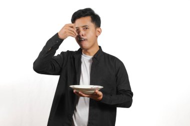 Siyah gömlekli genç Asyalı bir adam elinde kaşıkla tabaktan bir şey yiyor. Stüdyoda beyaz arka planda çekilmiş..