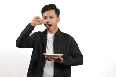 Siyah gömlekli genç Asyalı bir adam elinde kaşıkla tabaktan bir şey yiyor. Stüdyoda beyaz arka planda çekilmiş..