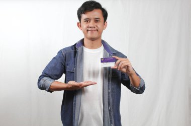 Mavi tişörtlü genç bir Asyalı adam beyaz bir arka planda izole edilmiş bir Endonezya akıllı kartını (KIP) tutarken ellerini kenarı işaret ediyor.