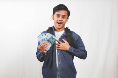 Sırt çantalı, elinde para tutan ve heyecanlı görünen mutlu, genç bir adam. Kot gömlek giyiyor ve beyaz bir arka planı olan parlak bir stüdyo ortamında duruyor..