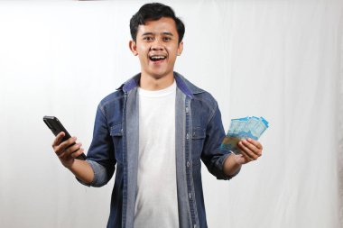 Gündelik giysiler içinde gülümseyen genç bir adam bir elinde akıllı bir telefon, diğer elinde ise bir yığın Endonezya Rupia banknotları tutuyor. Olumlu ve kutlama jestleri sergiliyor..