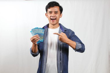 Sıradan bir kot gömlek giyen neşeli genç adam elinde bir yığın Endonezya Rupiah banknotu tutarken geniş bir gülümseme sergiliyor. Parayı mutlu bir ifadeyle gösteriyor, başarı ve zenginlik öneriyor..