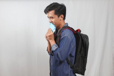 Kot ceketli genç Asyalı adam, siyah bir sırt çantası taşıyor, yüzünü birkaç banknotla kapatmaktan çok utanıyor. Düz beyaz bir arkaplanda kenar açıyla görülüyor.