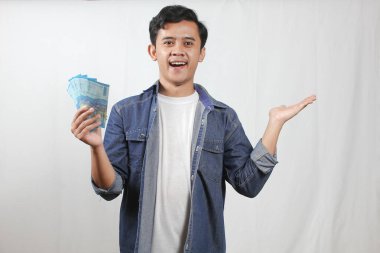 Kot gömlekli neşeli genç bir adam bir elinde temiz beyaz bir arka planda el kol hareketi yaparken diğer elinde bir yığın Endonezya Rupiah parası sunuyor.