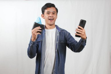 Kot gömlek giyen Asyalı adam elinde akıllı telefon ve cüzdanda beyaz arka planda izole edilmiş mutlu bir yüz ifadesi gösteren rupiah banknotları tutuyor.