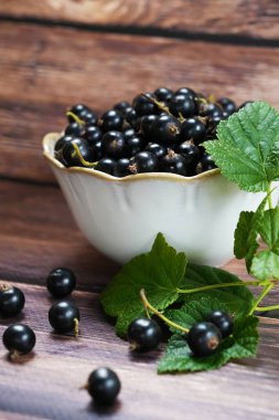 Yeşil yapraklar ve dağınık kuşüzümü ile çevrili ahşap bir yüzeyde taze kuşüzümü ile dolu beyaz bir kase..