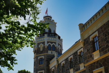 Ağaçlarla çevrili, yüksek kulesi ve bayrağı olan tarihi bir taş bina..