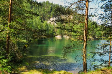 Bereketli yeşil ağaçlar ve açık mavi gökyüzünün altındaki kayalık uçurumlarla çevrili sakin bir orman gölü..