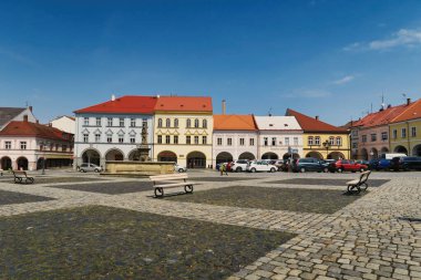Renkli tarihi binaları, kaldırımları ve bankları olan pitoresk bir kasaba meydanı. Gökyüzü açık ve mavi ve cadde boyunca birçok araba park halinde..