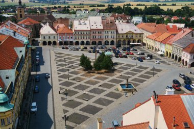 Ağaçlarla çevrili, renkli cepheli ve kırmızı çatıları olan tarihi binalarla çevrili bir şehir meydanı manzarası. Arabalar meydanda park halinde ve manzarada arka planda tarlalar var..