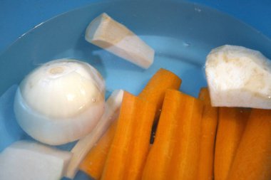 Havuç, maydanoz kökü, kohlrabi, kereviz kökü ve suyla dolu mavi kasede soğan da dahil olmak üzere taze soyulmuş sebzeler..