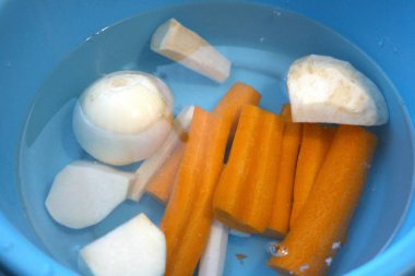 Havuç, maydanoz kökü, kohlrabi, kereviz kökü ve suyla dolu mavi kasede soğan da dahil olmak üzere taze soyulmuş sebzeler..