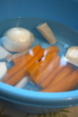 Havuç, maydanoz kökü, kohlrabi, kereviz kökü ve suyla dolu mavi kasede soğan da dahil olmak üzere taze soyulmuş sebzeler..