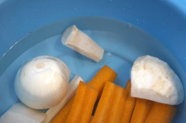 Havuç, maydanoz kökü, kohlrabi, kereviz kökü ve suyla dolu mavi kasede soğan da dahil olmak üzere taze soyulmuş sebzeler..