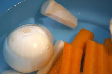 Havuç, maydanoz kökü, kohlrabi, kereviz kökü ve suyla dolu mavi kasede soğan da dahil olmak üzere taze soyulmuş sebzeler..