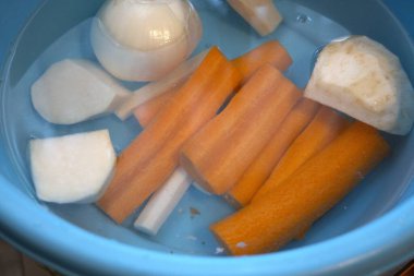 Havuç, maydanoz kökü, kohlrabi, kereviz kökü ve suyla dolu mavi kasede soğan da dahil olmak üzere taze soyulmuş sebzeler..