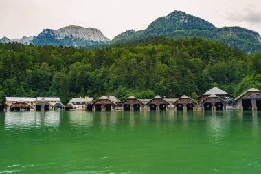 Vintage wooden boathouses line the calm, green waters, surrounded by dense trees and towering mountains under a cloudy sky, creating a tranquil atmosphere in nature. Konigssee lake Bavaria German Alps clipart