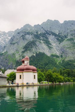 A white church with red domes stands by the tranquil waters of a lake, surrounded by lush greenery and majestic mountains on a sunny day. Konigssee lake Bavaria German Alps Berchtesgaden church by the clipart