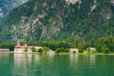 A white church with red domes stands by the tranquil waters of a lake, surrounded by lush greenery and majestic mountains on a sunny day. Konigssee lake Bavaria German Alps Berchtesgaden church by the clipart