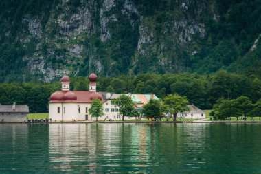 Güneşli bir günde, yemyeşil ve görkemli dağlarla çevrili, kırmızı kubbeleri olan beyaz bir kilise gölün sakin sularının yanında duruyor. Konigssee Gölü Bavyera Alman Alpleri Berchtesgaden Kilisesi